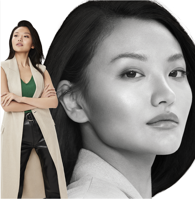 A woman in a long beige vest, green top, and black pants, standing with arms crossed, looking poised